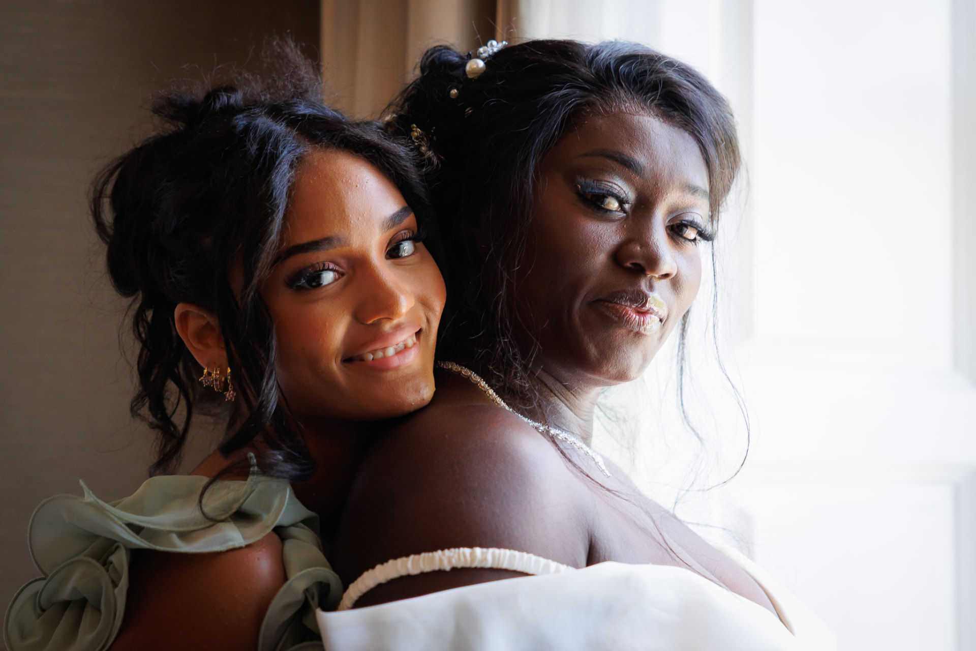 Bride and bridesmaid cheek to cheek in soft window light before the ceremony